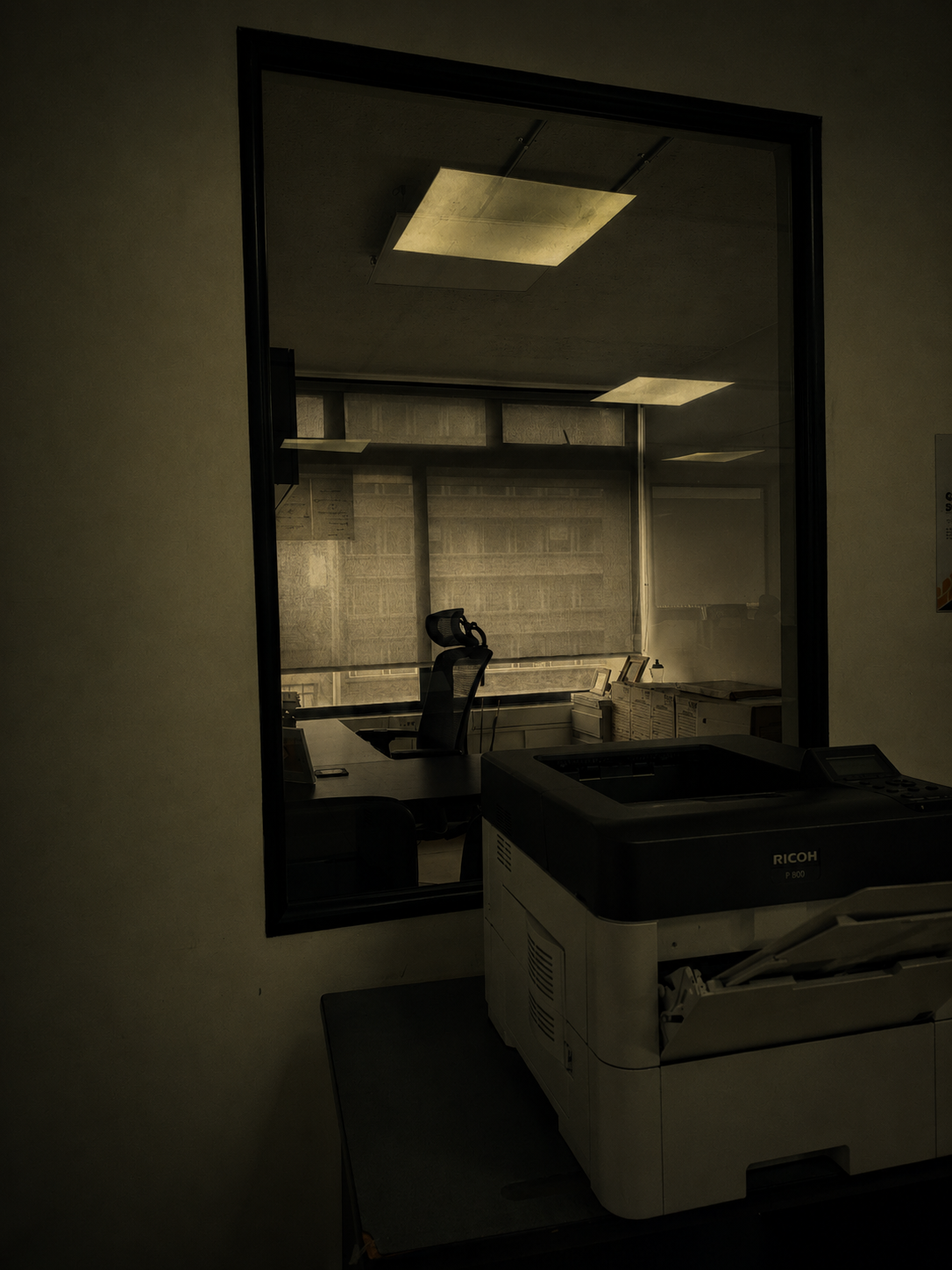 Ventana hacia una oficina vacía con una impresora en primer plano y luz cenital cayendo sobre un escritorio desocupado.